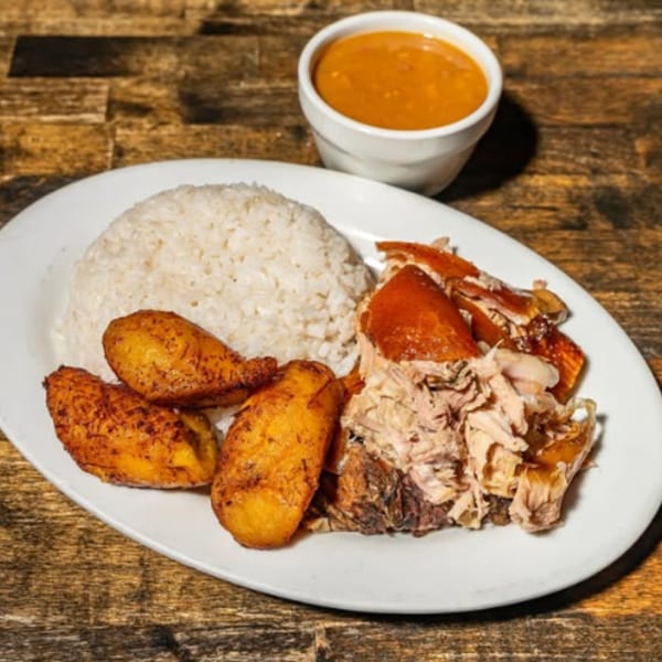 Cuban roasted pork with crispy skin, white rice, fried plantains, and a side of orange sauce.