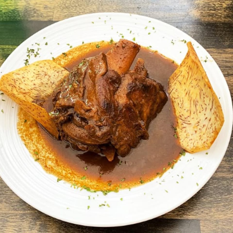 Cuban-style braised pork shank in a rich brown sauce, garnished with parsley and two taro chips.
